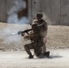Two military personnel in desert camouflage uniforms are firing a shoulder-launched rocket or recoilless weapon system, with visible backblast smoke and dust cloud generated by the discharge. One soldier is in a kneeling position operating the weapon while a second stands directly behind, both wearing combat helmets and tactical gear. The setting appears to be a training range or arid operational environment with a concrete barrier wall in the background.