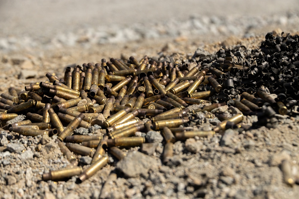 The image shows a large pile of spent brass cartridge casings scattered on sandy, gravelly ground. To the right, there is a separate pile of dark, fragmented metallic debris, likely shrapnel or deformed bullet fragments. The scene is consistent with a shooting range or post-firing cleanup area.