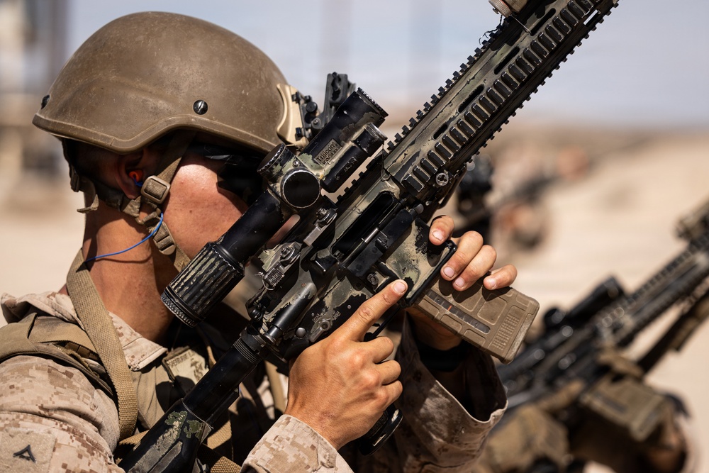 A military service member in desert camouflage uniform and tactical helmet is aiming a rifle equipped with an optical scope and accessories. The image is a close-up shot emphasizing the weapon and the shooter's stance. Additional personnel with weapons are visible in the blurred background, suggesting a training exercise or operational environment.