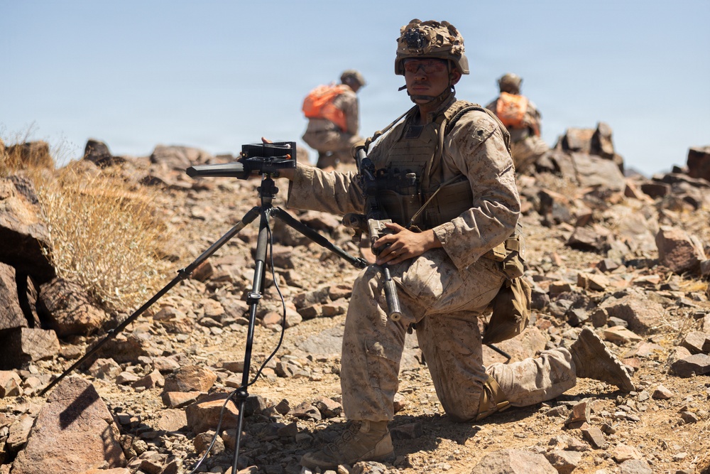 A military service member in desert camouflage uniform is kneeling on rocky terrain while operating a tripod-mounted electronic device, likely a targeting or communications system. The arid, rocky landscape and clear blue sky suggest a desert training environment. Two additional personnel wearing orange safety vests are visible in the background.