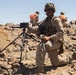 A military service member in desert camouflage uniform is kneeling on rocky terrain while operating a tripod-mounted electronic device, likely a targeting or communications system. The arid, rocky landscape and clear blue sky suggest a desert training environment. Two additional personnel wearing orange safety vests are visible in the background.