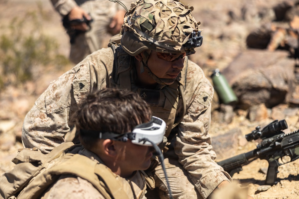 Two military personnel in desert camouflage uniforms are engaged in what appears to be a field training exercise in an arid, rocky environment. One soldier wearing a camouflage-netted helmet with a night vision mount is leaning over a second soldier who is wearing FPV (first-person view) drone goggles connected by a cable. A rifle with an optic scope is visible in the foreground right.