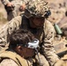 Two military personnel in desert camouflage uniforms are engaged in what appears to be a field training exercise in an arid, rocky environment. One soldier wearing a camouflage-netted helmet with a night vision mount is leaning over a second soldier who is wearing FPV (first-person view) drone goggles connected by a cable. A rifle with an optic scope is visible in the foreground right.