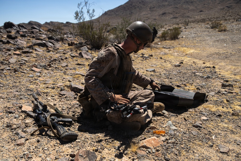 A military service member in desert camouflage uniform and helmet is seated on rocky ground in an arid desert environment, appearing to assemble or prepare a small unmanned aerial vehicle (drone). Their personal rifle is laid on the ground nearby. The setting appears to be a training exercise based on the controlled environment and desert terrain.