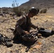 A military service member in desert camouflage uniform and helmet is seated on rocky ground in an arid desert environment, appearing to assemble or prepare a small unmanned aerial vehicle (drone). Their personal rifle is laid on the ground nearby. The setting appears to be a training exercise based on the controlled environment and desert terrain.