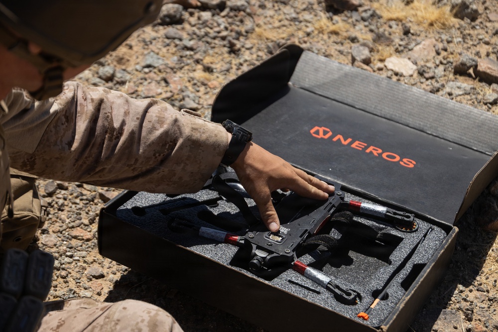 A person wearing desert camouflage military uniform is handling equipment from a branded case labeled 'NEROS' in an arid, rocky outdoor environment. The case contains what appears to be a small drone or unmanned aerial vehicle (UAV) system nestled in custom foam padding. The setting and attire suggest a military field evaluation or deployment scenario.