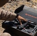 A person wearing desert camouflage military uniform is handling equipment from a branded case labeled 'NEROS' in an arid, rocky outdoor environment. The case contains what appears to be a small drone or unmanned aerial vehicle (UAV) system nestled in custom foam padding. The setting and attire suggest a military field evaluation or deployment scenario.