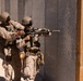 Two military personnel in desert camouflage uniforms are conducting what appears to be a close-quarters battle (CQB) training exercise near a simulated building entrance. One individual is aiming a rifle with an optic and suppressor toward a doorway, while the second provides guidance or support from behind. The setting is an arid, outdoor training environment with a constructed concrete wall and wooden door frame.