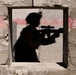 The image shows a shadow cast on a weathered concrete wall, depicting the silhouette of a person holding what appears to be a scoped rifle. The shadow is projected through or near a square window opening in the wall. A horizontal pink/red painted stripe runs across the wall surface.