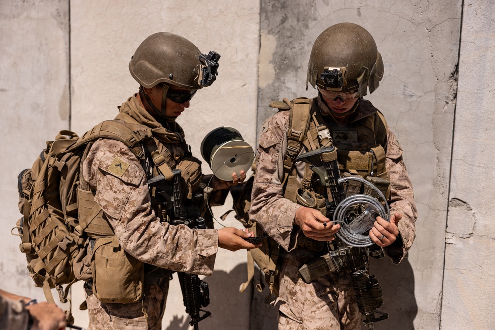 Two military personnel in desert camouflage uniforms are working with what appears to be demolition or communications equipment, including wire spools and coiled cable. They are positioned against a concrete wall in what appears to be a training or operational environment. Both soldiers are fully equipped with tactical gear, helmets with night vision mounts, body armor, and weapons.