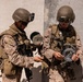 Two military personnel in desert camouflage uniforms are working with what appears to be demolition or communications equipment, including wire spools and coiled cable. They are positioned against a concrete wall in what appears to be a training or operational environment. Both soldiers are fully equipped with tactical gear, helmets with night vision mounts, body armor, and weapons.