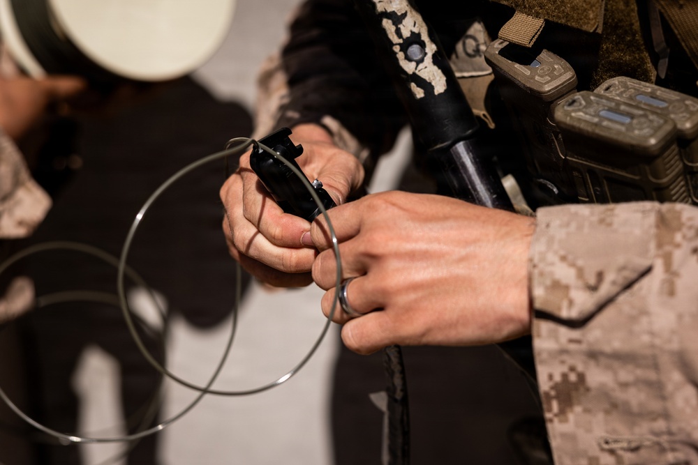 A person wearing military camouflage uniform (appears to be MARPAT desert pattern) is handling a black cylindrical device with a thin metal wire or cable coiled around it. Additional military equipment including what appears to be a rifle barrel and electronic devices are visible on their gear. The image is a close-up shot focused on the hands and equipment.