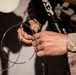 A person wearing military camouflage uniform (appears to be MARPAT desert pattern) is handling a black cylindrical device with a thin metal wire or cable coiled around it. Additional military equipment including what appears to be a rifle barrel and electronic devices are visible on their gear. The image is a close-up shot focused on the hands and equipment.
