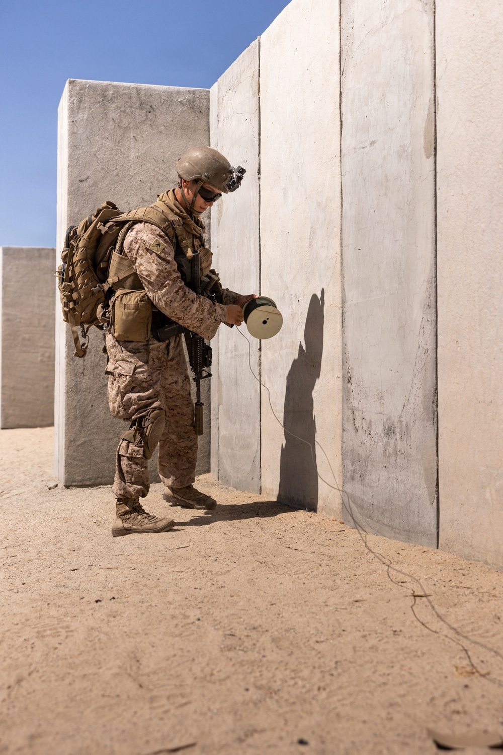 A U.S. Marine in MARPAT desert camouflage uniform is placing or handling a cylindrical device against a concrete wall in what appears to be a training environment. The soldier is fully equipped with tactical gear, helmet with NVG mount, body armor, and a slung rifle. The setting appears to be an arid, desert training range with concrete block structures.