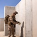 A U.S. Marine in MARPAT desert camouflage uniform is placing or handling a cylindrical device against a concrete wall in what appears to be a training environment. The soldier is fully equipped with tactical gear, helmet with NVG mount, body armor, and a slung rifle. The setting appears to be an arid, desert training range with concrete block structures.