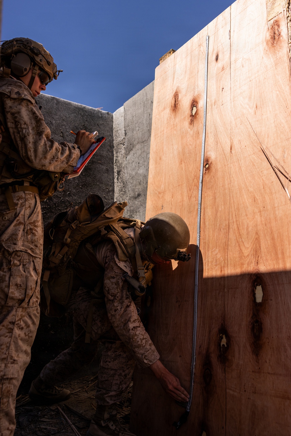 Two military personnel in desert camouflage uniforms are conducting a measurement activity near a large plywood panel and concrete barrier. One service member holds a red clipboard and appears to be recording data, while the other crouches at the base of the plywood holding a measuring tape extended vertically. The setting appears to be an outdoor training or assessment environment under clear blue skies.
