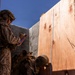 Two military personnel in desert camouflage uniforms are conducting a measurement activity near a large plywood panel and concrete barrier. One service member holds a red clipboard and appears to be recording data, while the other crouches at the base of the plywood holding a measuring tape extended vertically. The setting appears to be an outdoor training or assessment environment under clear blue skies.