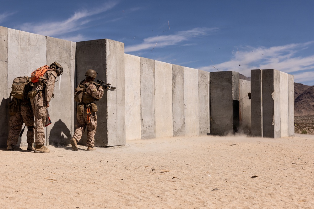 Two military personnel in desert camouflage uniforms are conducting a tactical training exercise at what appears to be a desert training facility. One soldier is aiming a rifle around a concrete wall while the other provides cover behind them. A breaching or explosive event appears to have just occurred at a concrete structure further down the range, evidenced by dust and debris visible near a doorway.