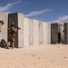 Two military personnel in desert camouflage uniforms are conducting a tactical training exercise at what appears to be a desert training facility. One soldier is aiming a rifle around a concrete wall while the other provides cover behind them. A breaching or explosive event appears to have just occurred at a concrete structure further down the range, evidenced by dust and debris visible near a doorway.