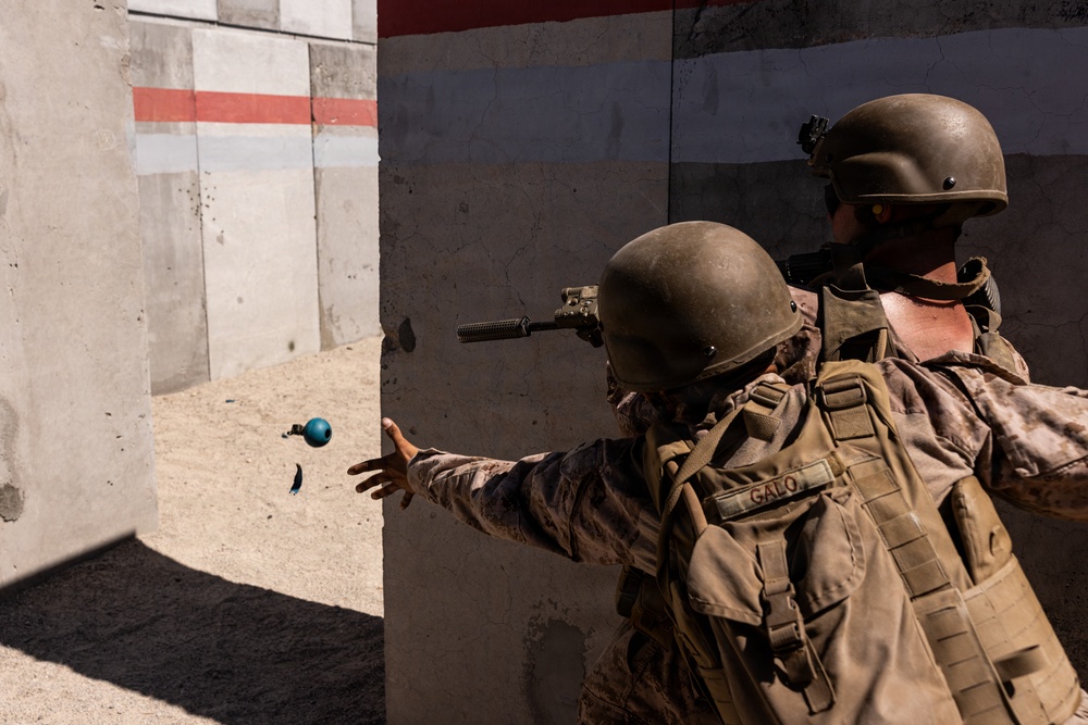 Two military personnel in full combat gear are conducting a training exercise near a concrete wall structure. The foreground soldier, identified by a name tape reading 'GALO', is throwing what appears to be a blue training grenade (simulated/inert) around a corner. Both soldiers are wearing helmets, body armor, and camouflage uniforms consistent with a military training environment.