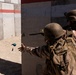 Two military personnel in full combat gear are conducting a training exercise near a concrete wall structure. The foreground soldier, identified by a name tape reading 'GALO', is throwing what appears to be a blue training grenade (simulated/inert) around a corner. Both soldiers are wearing helmets, body armor, and camouflage uniforms consistent with a military training environment.
