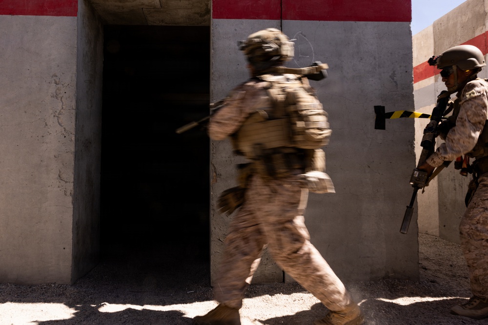 Two soldiers in desert camouflage uniforms and full tactical gear are conducting what appears to be a military training exercise involving building entry or clearing. One soldier is in motion entering a darkened doorway while a second soldier stands in a ready position nearby. The setting appears to be a purpose-built training facility with concrete walls painted with red and white markings.