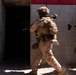 Two soldiers in desert camouflage uniforms and full tactical gear are conducting what appears to be a military training exercise involving building entry or clearing. One soldier is in motion entering a darkened doorway while a second soldier stands in a ready position nearby. The setting appears to be a purpose-built training facility with concrete walls painted with red and white markings.