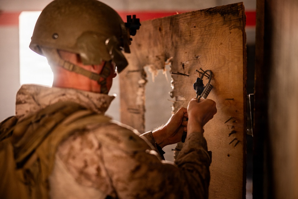 A military service member in desert camouflage uniform and combat helmet is working on a damaged wooden door or board, appearing to manipulate a lock or hardware component. The individual is wearing a face covering and has equipment mounted on their helmet. The setting appears to be an interior structure with visible damage to the wooden surface.