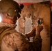 A military service member in desert camouflage uniform and combat helmet is working on a damaged wooden door or board, appearing to manipulate a lock or hardware component. The individual is wearing a face covering and has equipment mounted on their helmet. The setting appears to be an interior structure with visible damage to the wooden surface.
