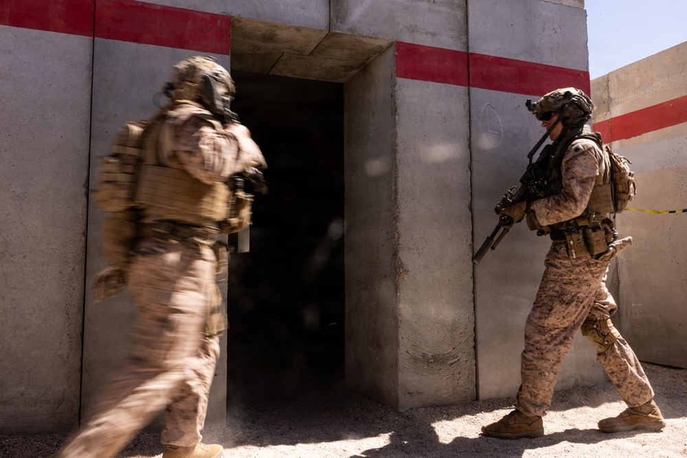 Two military personnel in full combat gear are conducting a building entry or clearing operation at what appears to be a training facility. One soldier is in motion entering a doorway (motion blur visible), while the second soldier stands in a ready position holding a rifle. The structure features concrete walls with red stripe markings, consistent with a military urban operations training environment.