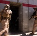 Two military personnel in full combat gear are conducting a building entry or clearing operation at what appears to be a training facility. One soldier is in motion entering a doorway (motion blur visible), while the second soldier stands in a ready position holding a rifle. The structure features concrete walls with red stripe markings, consistent with a military urban operations training environment.