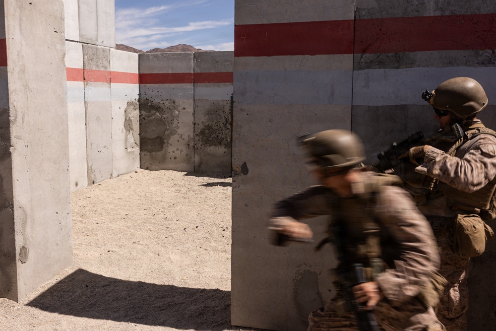 Two armed military personnel in desert camouflage uniforms and helmets are conducting a tactical training exercise at what appears to be a military training facility. One soldier is in motion (motion blur visible), moving through a gap in concrete walls, while a second soldier holds a covered position with a rifle raised. The concrete walls show evidence of previous live-fire training with visible impact marks and scorch patterns.