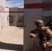 Two armed military personnel in desert camouflage uniforms and helmets are conducting a tactical training exercise at what appears to be a military training facility. One soldier is in motion (motion blur visible), moving through a gap in concrete walls, while a second soldier holds a covered position with a rifle raised. The concrete walls show evidence of previous live-fire training with visible impact marks and scorch patterns.