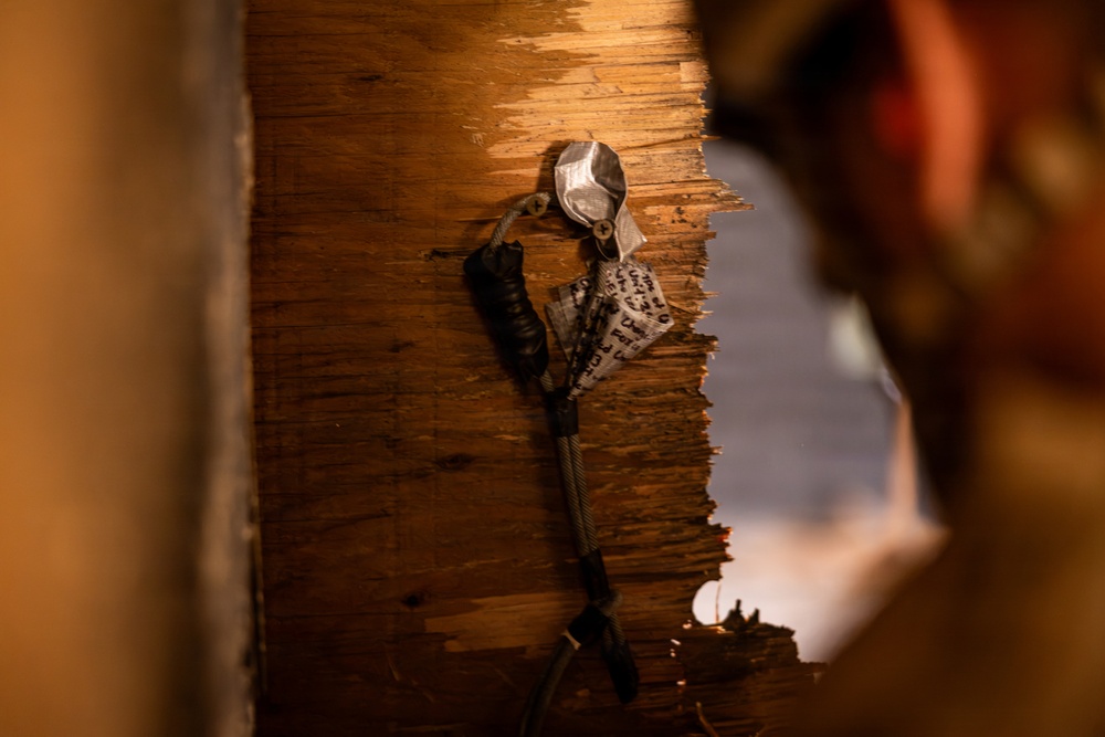 The image shows a close-up of a damaged or broken wooden wall or door frame with exposed splintered wood layers. Attached to the wood surface are cables or wires secured with what appears to be metal hardware and tape, along with a piece of paper or label with printed text. A person is partially visible and out of focus in the right foreground.