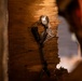 The image shows a close-up of a damaged or broken wooden wall or door frame with exposed splintered wood layers. Attached to the wood surface are cables or wires secured with what appears to be metal hardware and tape, along with a piece of paper or label with printed text. A person is partially visible and out of focus in the right foreground.
