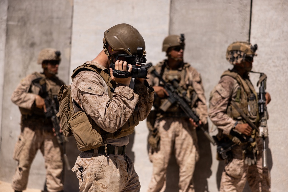 Four military personnel in desert camouflage (MARPAT pattern) are positioned against a concrete wall in what appears to be an arid environment. One soldier in the foreground is operating a Canon video/photo camera, while the remaining three carry assault rifles and appear to be providing security. The scene suggests a documentation or combat camera operation during a military exercise or deployment.