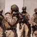 Four military personnel in desert camouflage (MARPAT pattern) are positioned against a concrete wall in what appears to be an arid environment. One soldier in the foreground is operating a Canon video/photo camera, while the remaining three carry assault rifles and appear to be providing security. The scene suggests a documentation or combat camera operation during a military exercise or deployment.