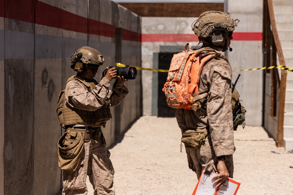 Two military personnel in desert camouflage uniforms are positioned in what appears to be a training or exercise environment. One soldier is photographing the other using a professional DSLR camera with a telephoto lens. The second individual wears a distinctive bright orange backpack with a visible number, suggesting a training exercise role identifier.