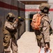 Two military personnel in desert camouflage uniforms are positioned in what appears to be a training or exercise environment. One soldier is photographing the other using a professional DSLR camera with a telephoto lens. The second individual wears a distinctive bright orange backpack with a visible number, suggesting a training exercise role identifier.