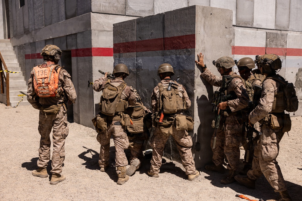 A group of military personnel in desert camouflage uniforms are stacked against a concrete wall during what appears to be a training exercise. The soldiers are positioned in a tactical formation near a building corner, suggesting a breach or room-clearing drill. The presence of a label '13E' on an orange backpack and safety tape in the background supports the training exercise context.