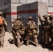 A group of military personnel in desert camouflage uniforms are stacked against a concrete wall during what appears to be a training exercise. The soldiers are positioned in a tactical formation near a building corner, suggesting a breach or room-clearing drill. The presence of a label '13E' on an orange backpack and safety tape in the background supports the training exercise context.