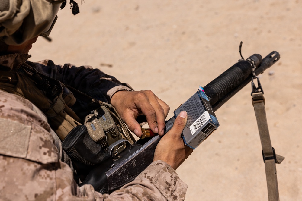 A military service member in desert camouflage (MARPAT pattern) is loading or handling ammunition from a commercial box into what appears to be a shotgun or grenade launcher attachment. The individual is equipped with tactical gear including pouches and a radio or communication device. The setting appears to be an arid, sandy outdoor training or operational environment.