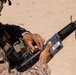 A military service member in desert camouflage (MARPAT pattern) is loading or handling ammunition from a commercial box into what appears to be a shotgun or grenade launcher attachment. The individual is equipped with tactical gear including pouches and a radio or communication device. The setting appears to be an arid, sandy outdoor training or operational environment.