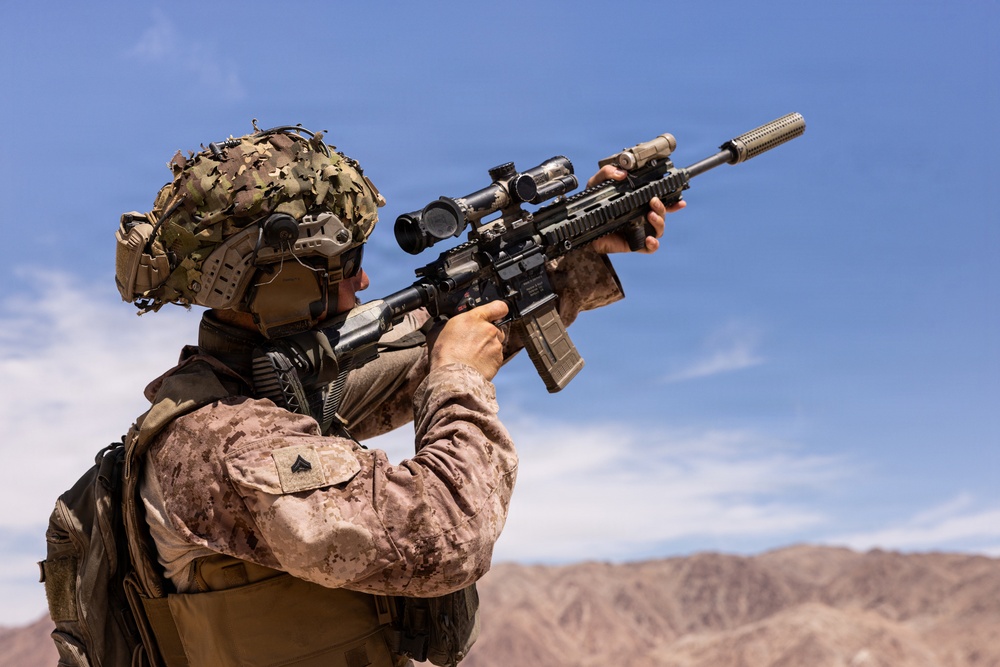 A military service member in desert camouflage (MARPAT pattern) is shown aiming a scoped assault rifle in an arid, mountainous environment. The soldier is equipped with full tactical gear including a helmet with camouflage netting and communications equipment. The image appears to depict a combat or training scenario in a desert region.