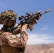 A military service member in desert camouflage (MARPAT pattern) is shown aiming a scoped assault rifle in an arid, mountainous environment. The soldier is equipped with full tactical gear including a helmet with camouflage netting and communications equipment. The image appears to depict a combat or training scenario in a desert region.