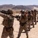 A line of military personnel in desert camouflage uniforms are conducting a live-fire or training exercise in an arid desert environment. The soldiers are positioned in a firing line, aiming rifles in the same direction. The setting appears to be a designated military training range based on the organized formation and safety equipment visible.