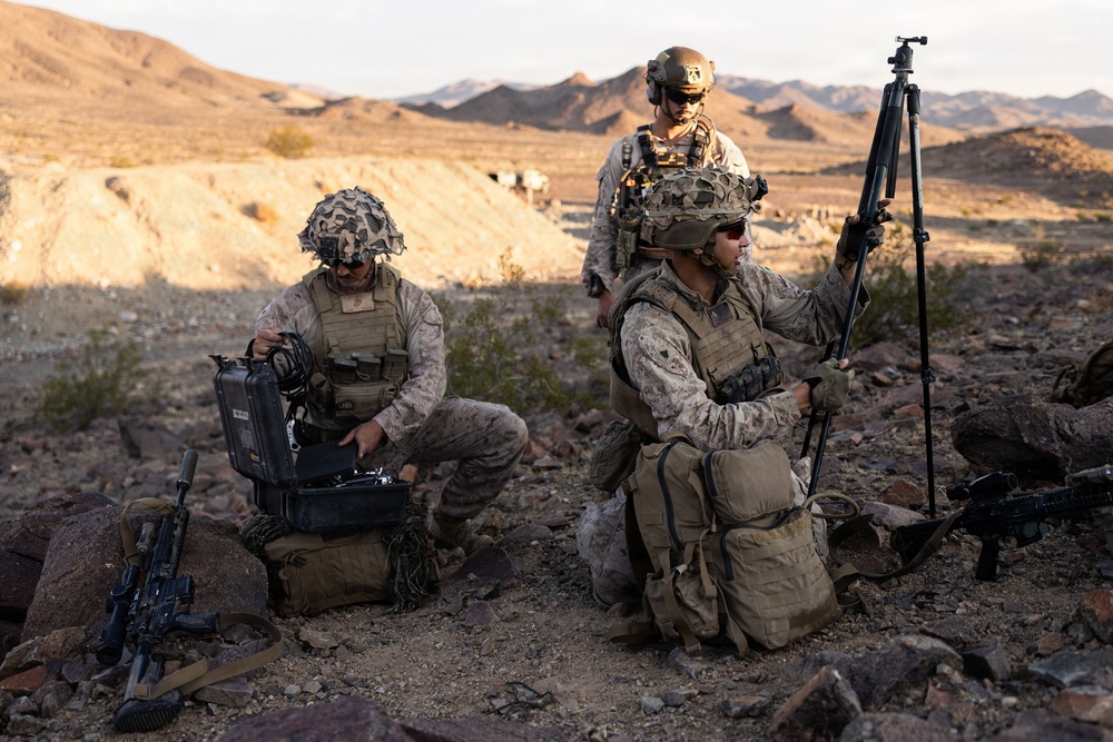 Three military personnel in desert camouflage uniforms are operating equipment in an arid, rocky terrain with mountains visible in the background. Two soldiers are kneeling while one operates what appears to be a ruggedized equipment case and another holds a tripod-mounted device. The setting and uniforms are consistent with a U.S. Marine Corps field training exercise in a desert environment.
