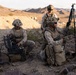 Three military personnel in desert camouflage uniforms are operating equipment in an arid, rocky terrain with mountains visible in the background. Two soldiers are kneeling while one operates what appears to be a ruggedized equipment case and another holds a tripod-mounted device. The setting and uniforms are consistent with a U.S. Marine Corps field training exercise in a desert environment.