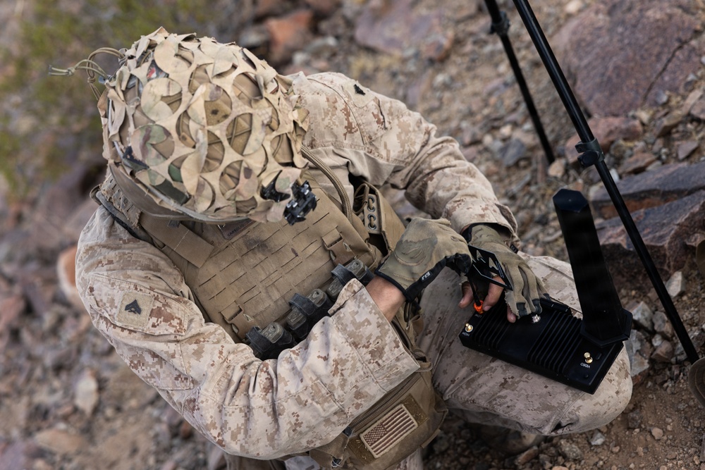 A military personnel wearing desert MARPAT camouflage uniform and a camouflage-netted helmet is crouched on rocky terrain while operating a black electronic device. The soldier is equipped with full tactical gear including a plate carrier vest with MOLLE webbing and fingerless tactical gloves. A tripod-mounted antenna or sensor system is visible in the background to the right.