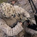 A military personnel wearing desert MARPAT camouflage uniform and a camouflage-netted helmet is crouched on rocky terrain while operating a black electronic device. The soldier is equipped with full tactical gear including a plate carrier vest with MOLLE webbing and fingerless tactical gloves. A tripod-mounted antenna or sensor system is visible in the background to the right.